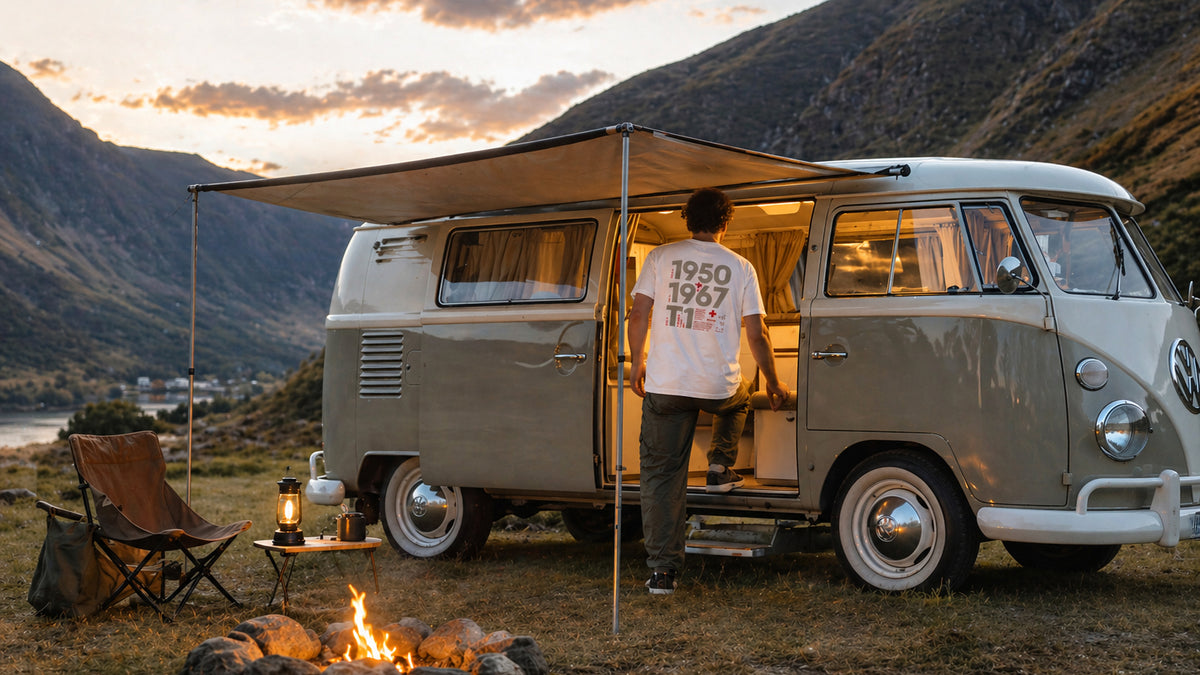 Person wearing the Vanture T1 Era T-Shirt beside a vintage VW T1 campervan at a mountain campsite during sunset.